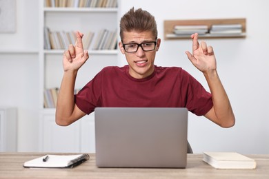 Worried student with crossed fingers at table with laptop indoors. Hope for good exam result