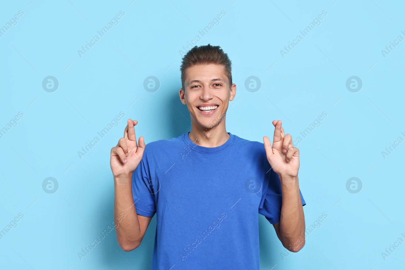 Photo of Happy student with crossed fingers on light blue background. Hope for good exam result