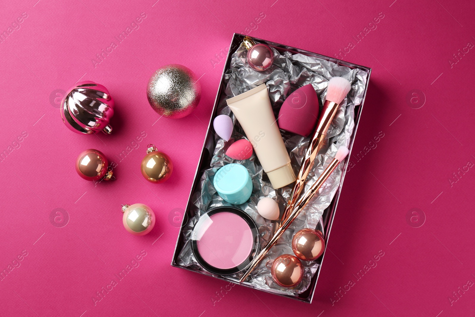 Photo of Christmas present. Decorative cosmetics in gift box and festive balls on dark pink background, top view