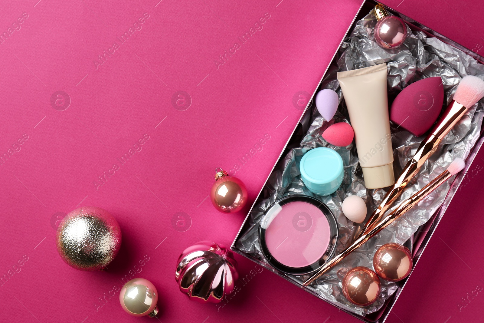 Photo of Christmas present. Decorative cosmetics in gift box and festive balls on dark pink background, top view. Space for text