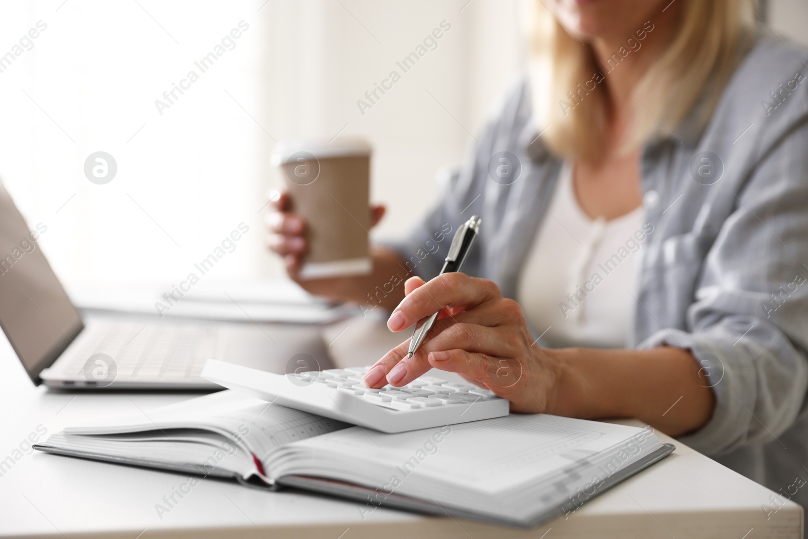 Photo of Budget planning. Woman with paper cup of coffee using calculator at table indoors, closeup