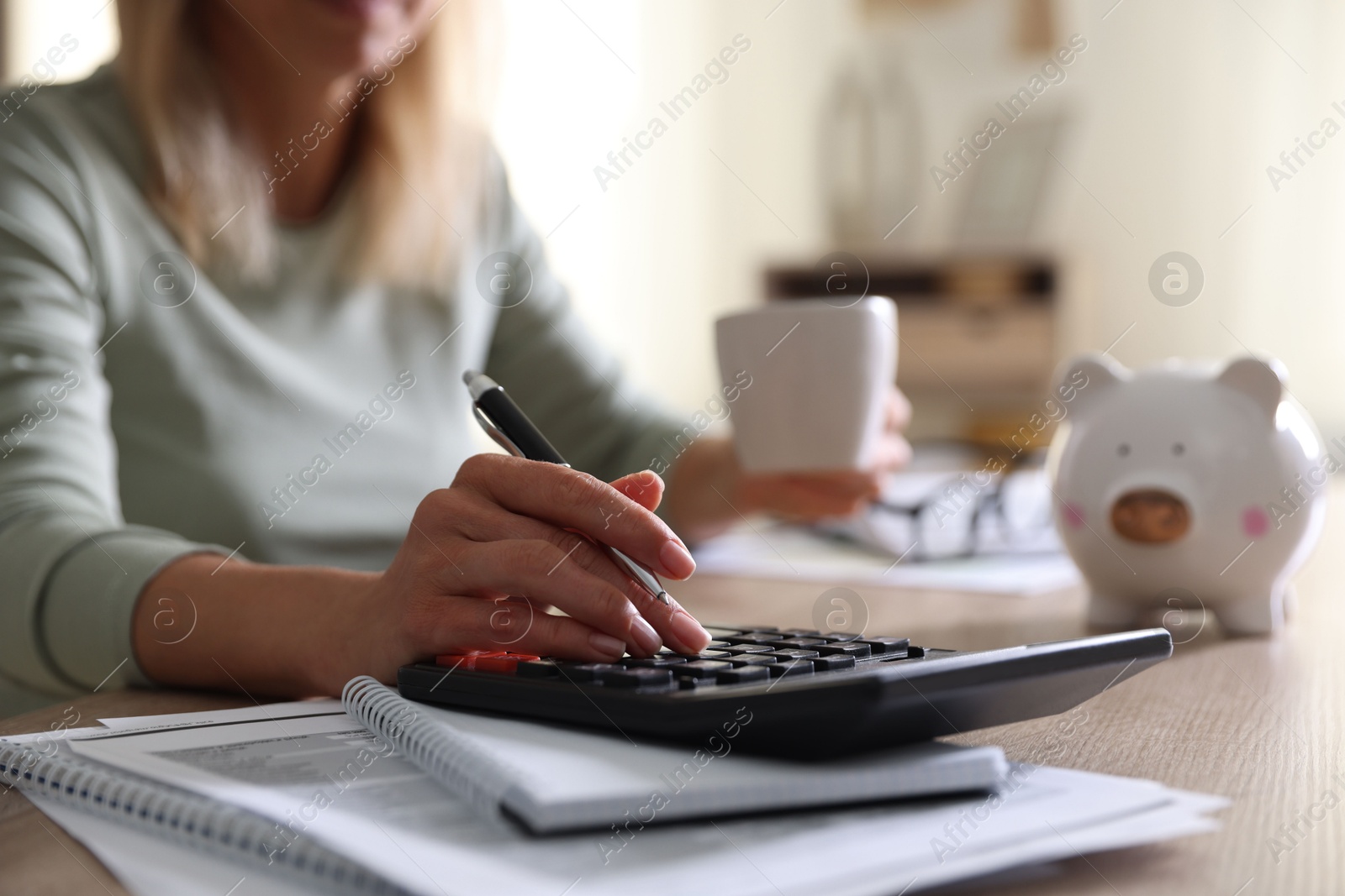 Photo of Budget planning. Woman with cup of drink using calculator at table indoors, closeup