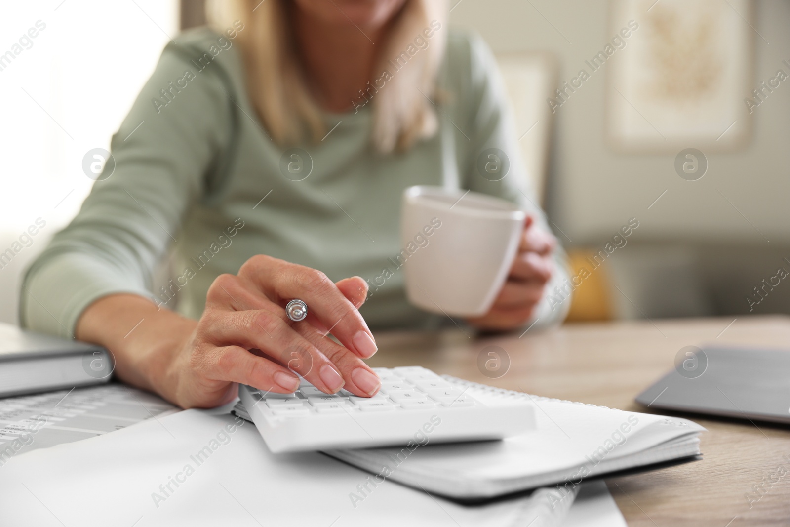 Photo of Budget planning. Woman with cup of drink using calculator at table indoors, closeup