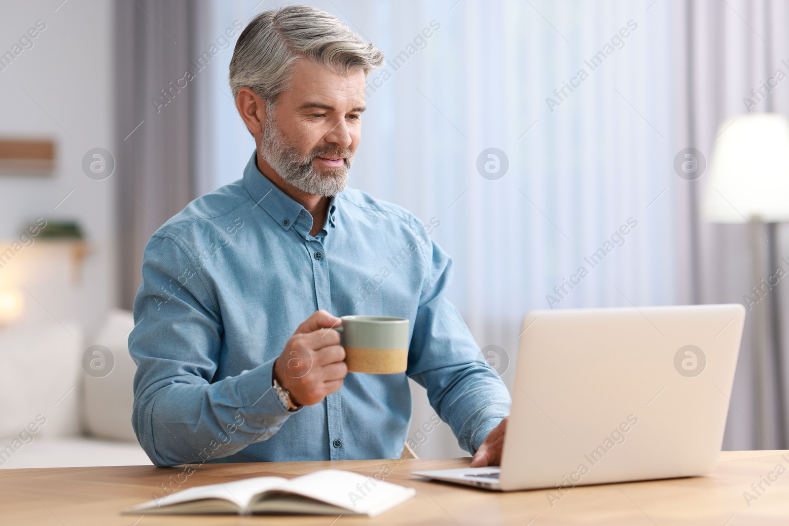 Photo of Middle aged man with cup of drink working with laptop at table indoors