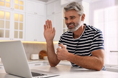 Photo of Happy middle aged man with cup of drink having video chat via laptop at white marble table in kitchen