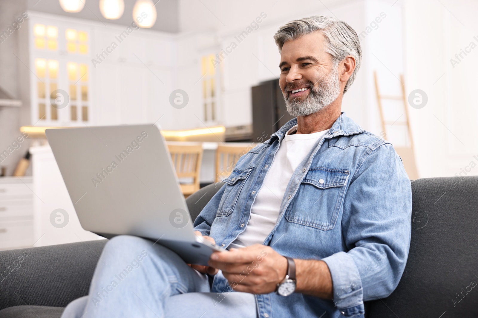 Photo of Happy middle aged man using laptop on sofa in kitchen