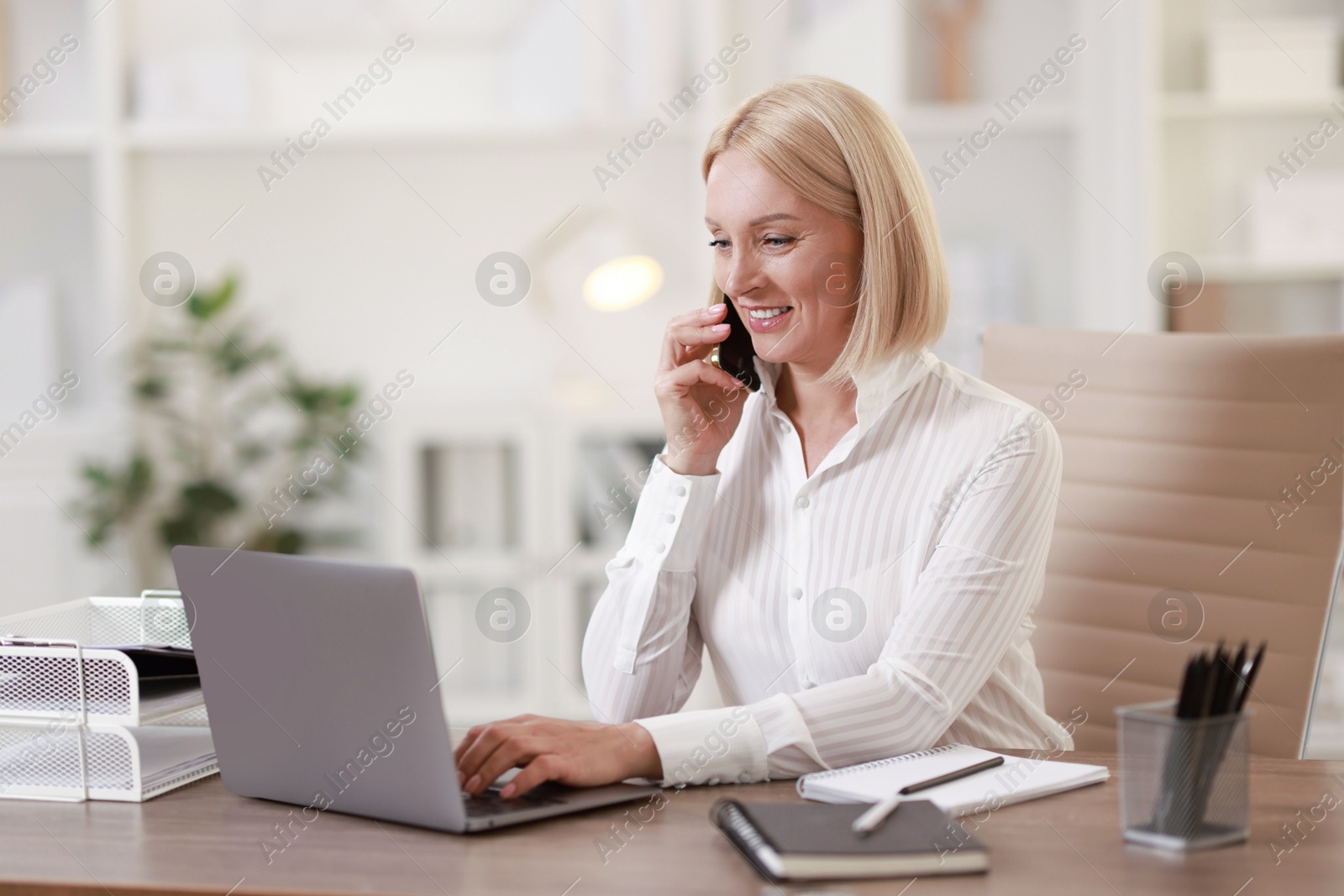 Photo of Smiling middle aged woman talking by smartphone at table in office