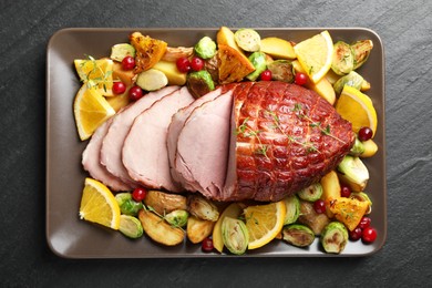 Tasty baked ham served with vegetables and oranges on black table, top view