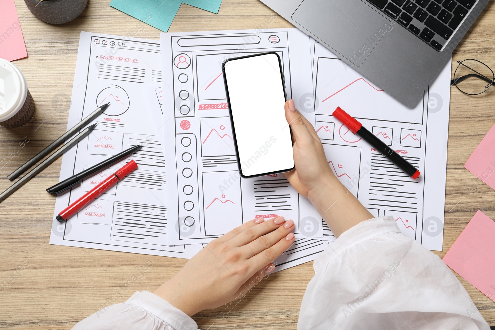 Photo of Website designer holding smartphone at wooden table with wireframes and stationery, top view