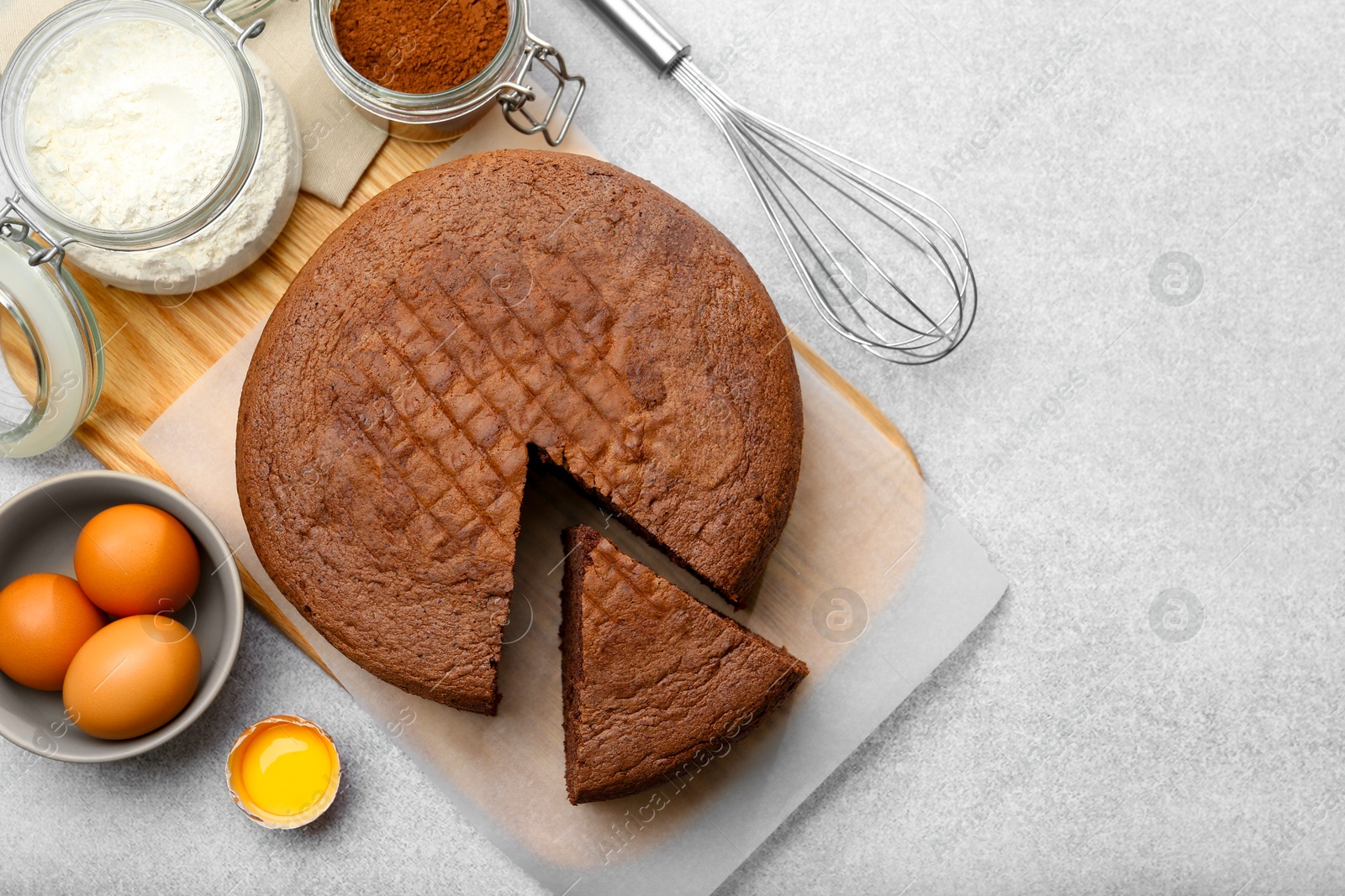 Photo of Cut chocolate sponge cake, whisk and ingredients on light table, flat lay