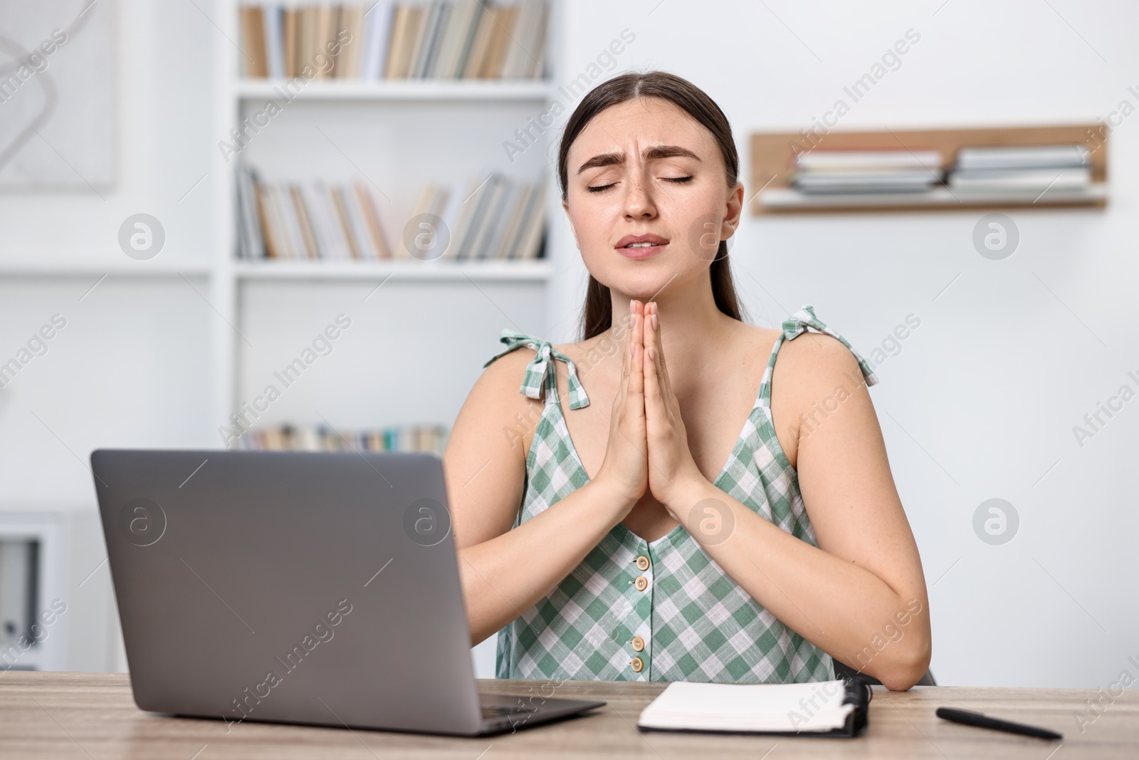 Photo of Worried student praying for good exam result at table with laptop indoors