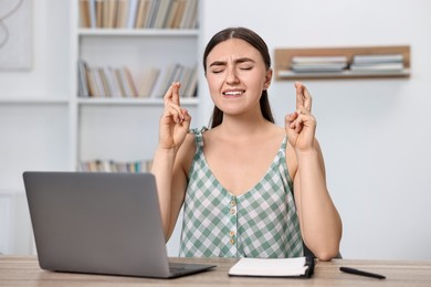 Photo of Worried student with crossed fingers at table with laptop indoors. Hope for good exam result