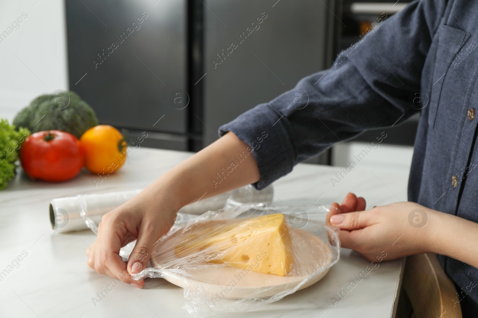 Photo of Woman putting plastic food wrap over plate with cheese at white marble table in kitchen, closeup