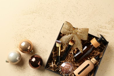 Photo of Christmas gift box with cosmetic products and festive balls on beige background, flat lay. Space for text