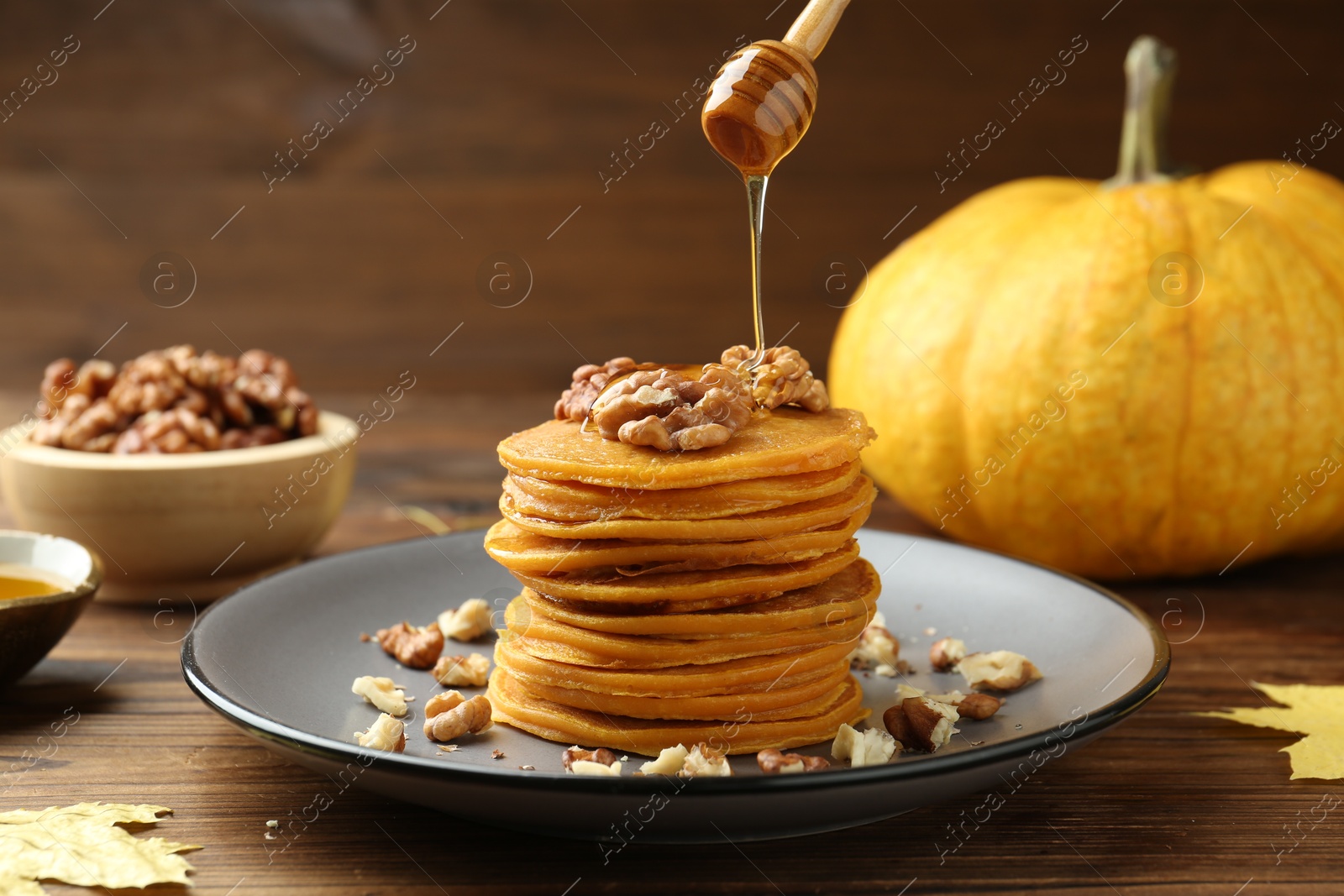 Photo of Pouring honey onto tasty pumpkin pancakes with walnuts at wooden table, closeup