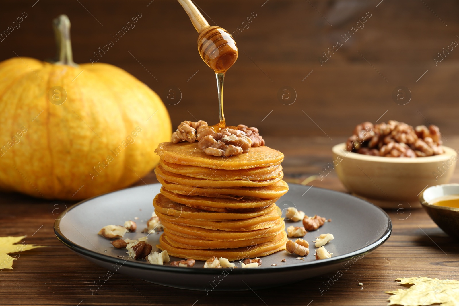 Photo of Pouring honey onto tasty pumpkin pancakes with walnuts at wooden table, closeup