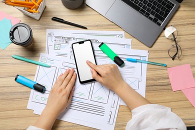 Photo of Website designer using smartphone at wooden table with wireframes and laptop, top view