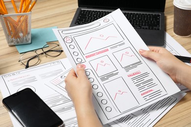 Photo of Website designer holding wireframe at wooden table with smartphone and laptop, closeup