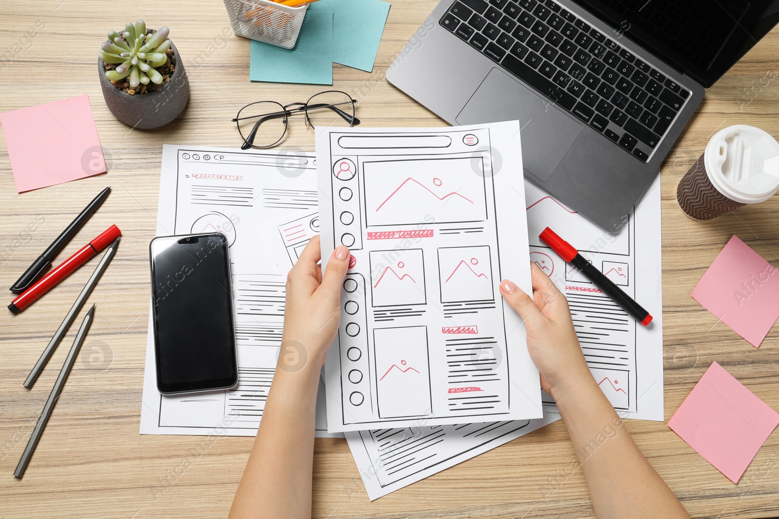 Photo of Website designer holding wireframe at wooden table with stationery and laptop, top view