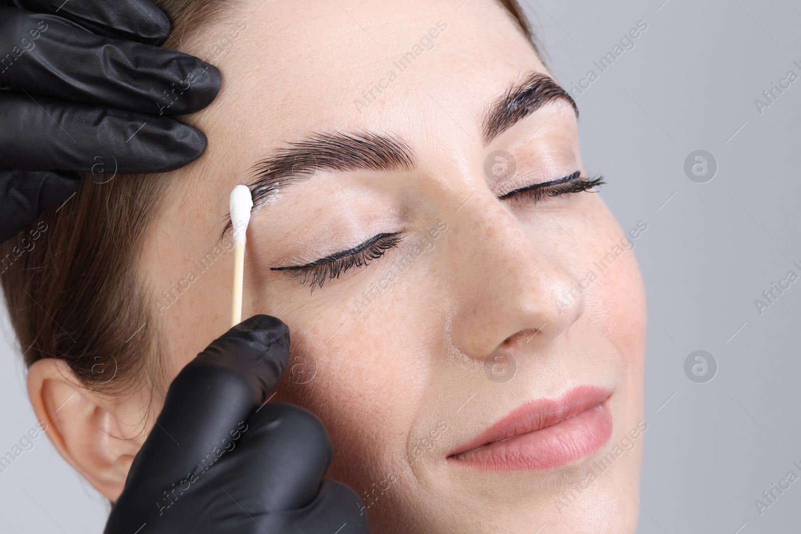 Photo of Brow lamination. Cosmetologist applying cream onto woman's eyebrows against grey background, closeup