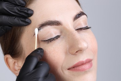 Photo of Brow lamination. Cosmetologist applying cream onto woman's eyebrows against grey background, closeup