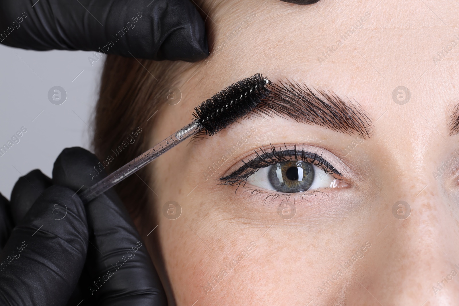 Photo of Brow lamination. Cosmetologist combing woman's eyebrows with brush against grey background, closeup