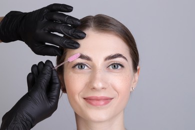 Photo of Brow lamination. Cosmetologist combing woman's eyebrows with brush against grey background, closeup