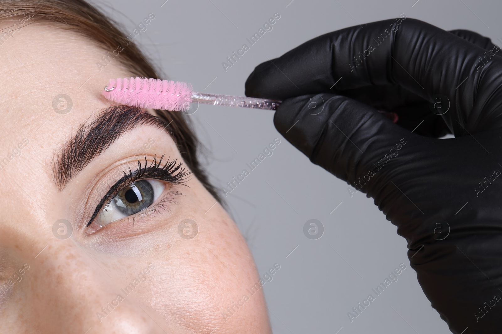 Photo of Brow lamination. Cosmetologist combing woman's eyebrows with brush against grey background, closeup