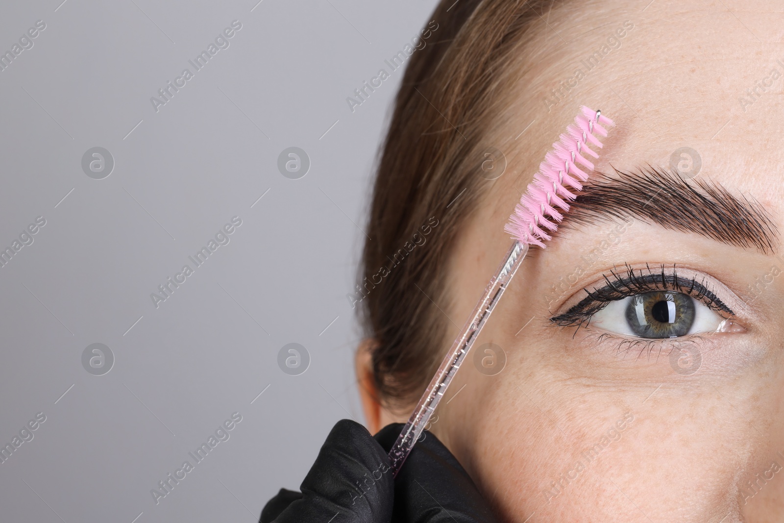 Photo of Brow lamination. Cosmetologist combing woman's eyebrows with brush against grey background, closeup. Space for text