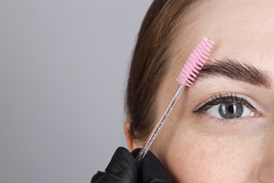 Brow lamination. Cosmetologist combing woman's eyebrows with brush against grey background, closeup. Space for text