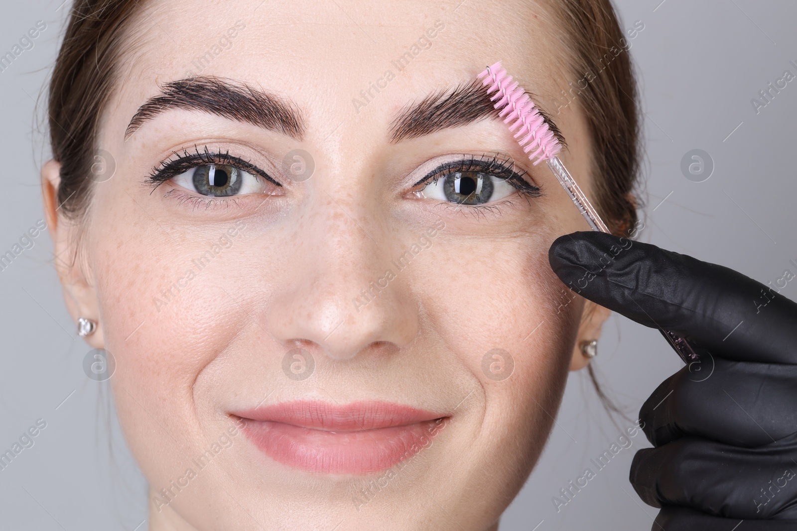 Photo of Brow lamination. Cosmetologist combing woman's eyebrows with brush against grey background, closeup