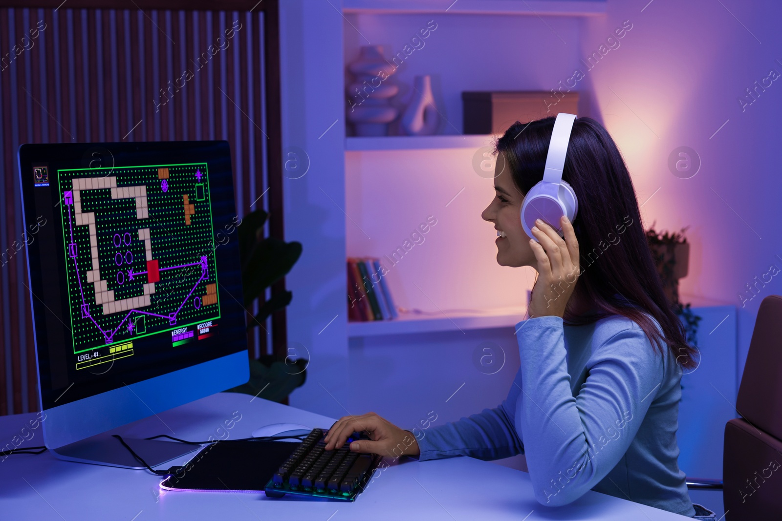 Photo of Smiling woman in headphones playing video game with keyboard at table indoors