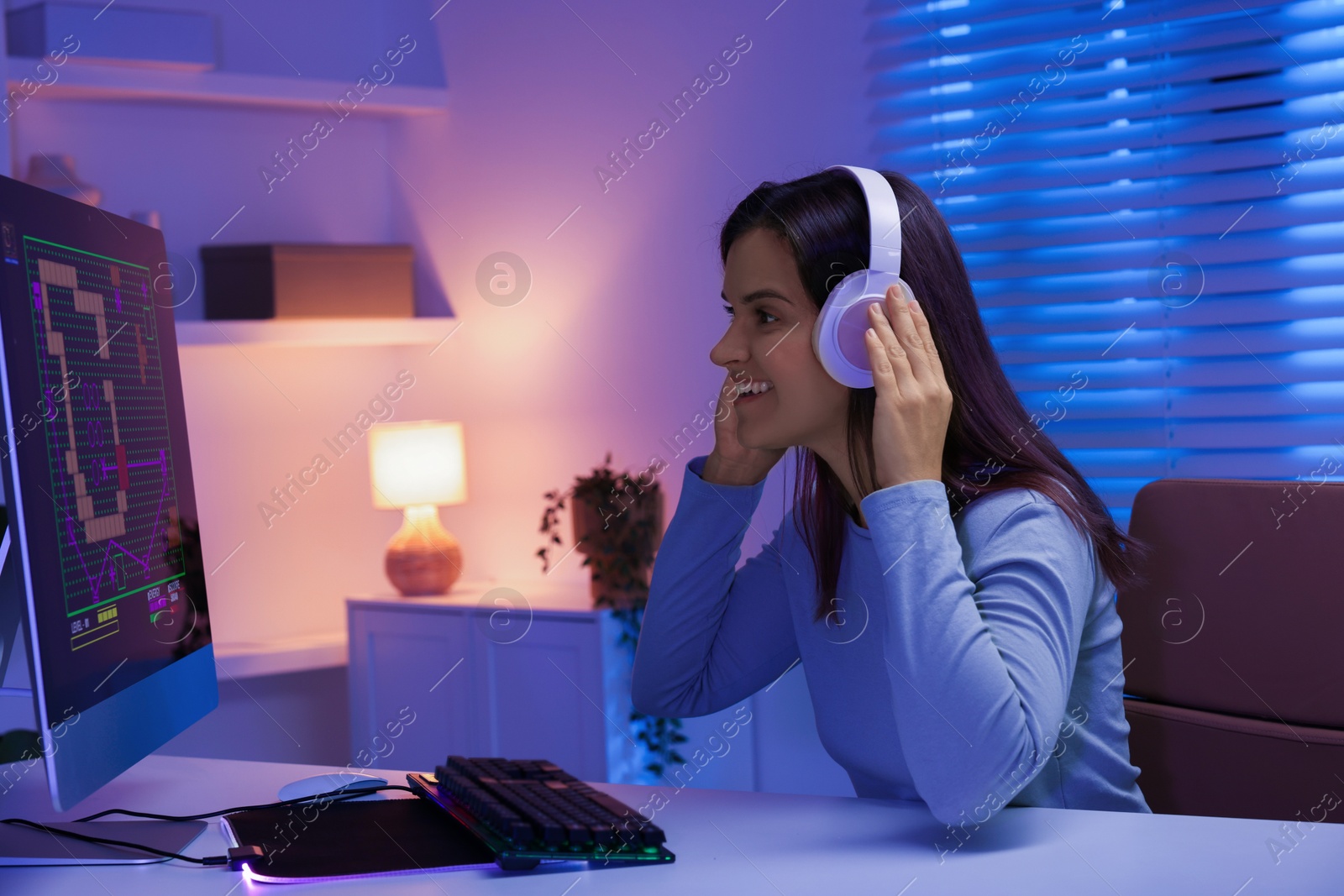 Photo of Smiling woman in headphones playing video game with keyboard at table indoors