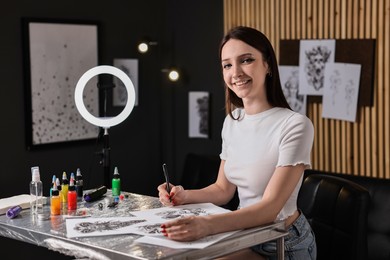 Photo of Smiling tattoo artist drawing sketch at table with supplies in salon