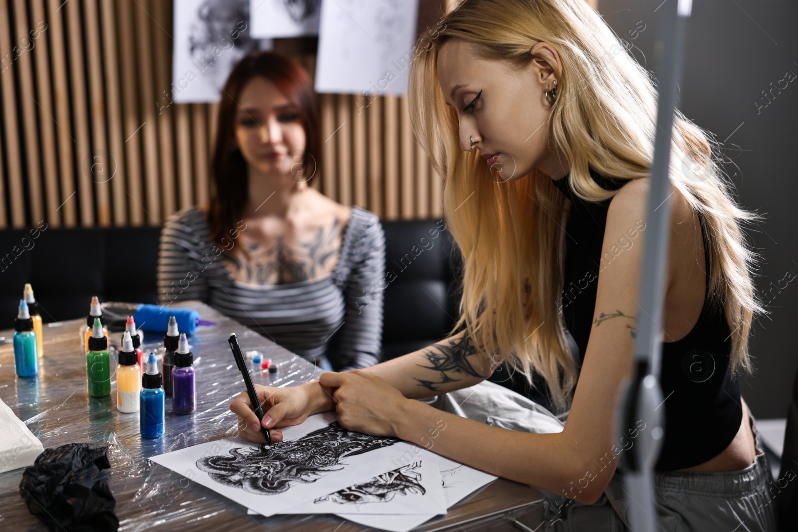 Photo of Tattoo artist drawing sketch at table with supplies near client in salon, selective focus