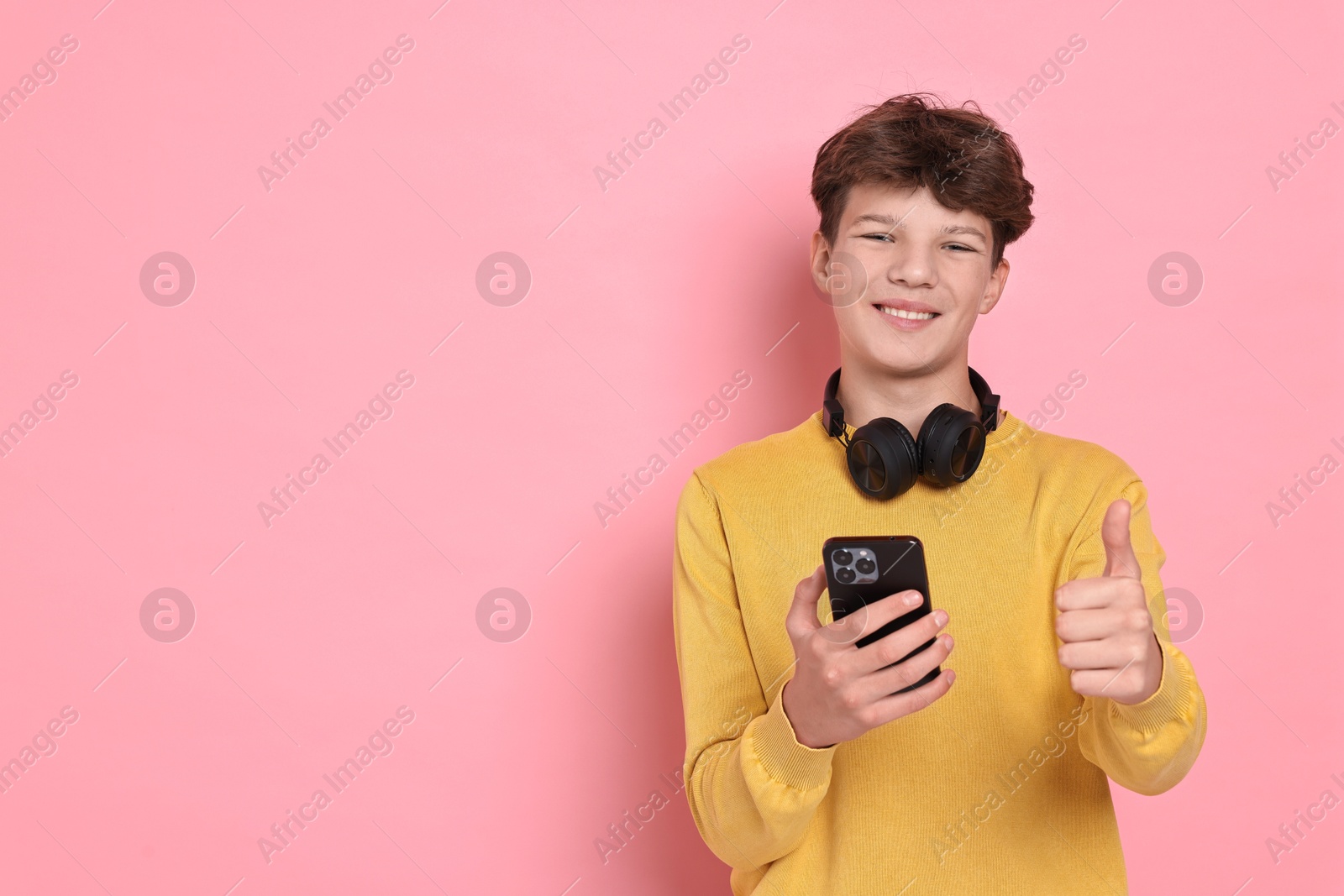 Photo of Teenage boy with headphones and smartphone showing thumbs up on pink background, space for text