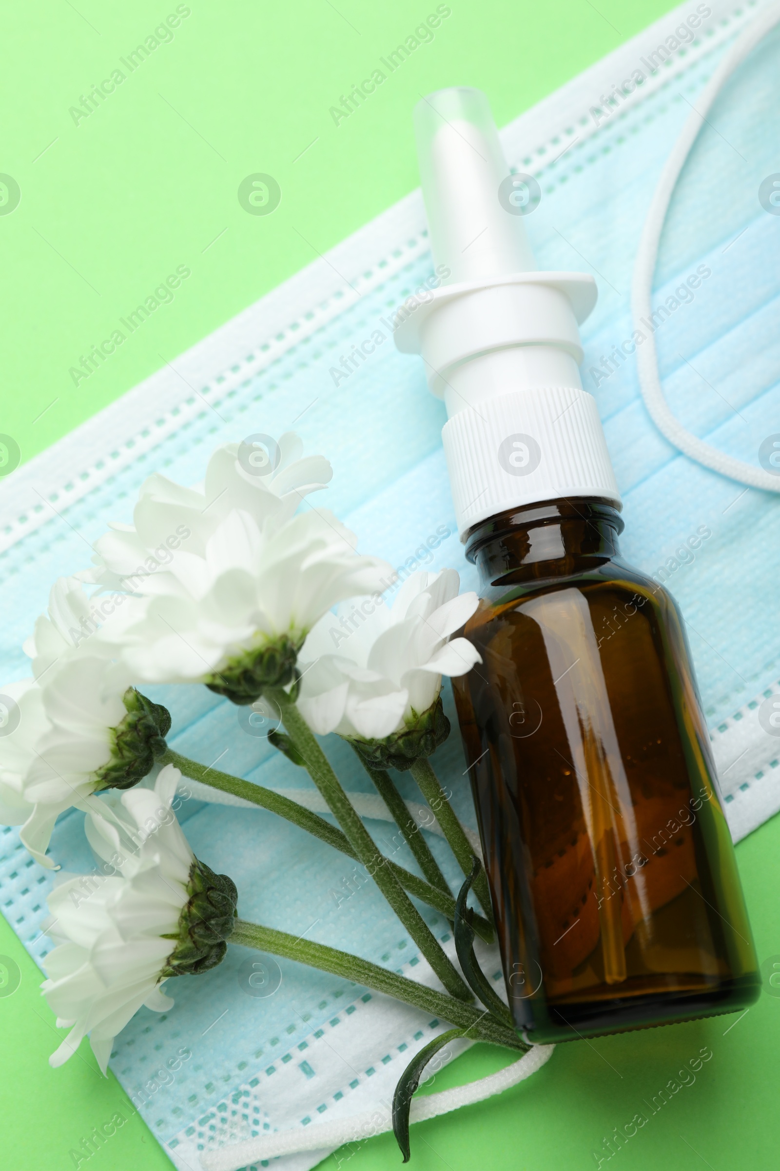 Photo of Allergy treatment. Nasal spray, protective mask and flowers on green background, top view