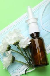 Photo of Allergy treatment. Nasal spray, protective mask and flowers on green background, top view