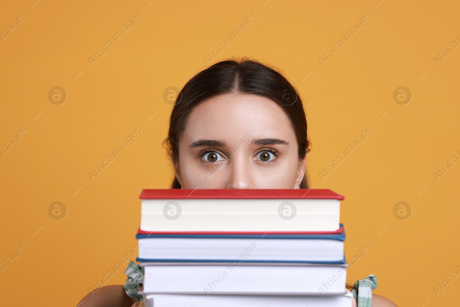 Photo of Stressful student with stack of books before exam on orange background, closeup