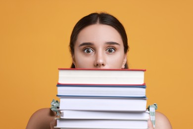 Stressful student with stack of books before exam on orange background, closeup