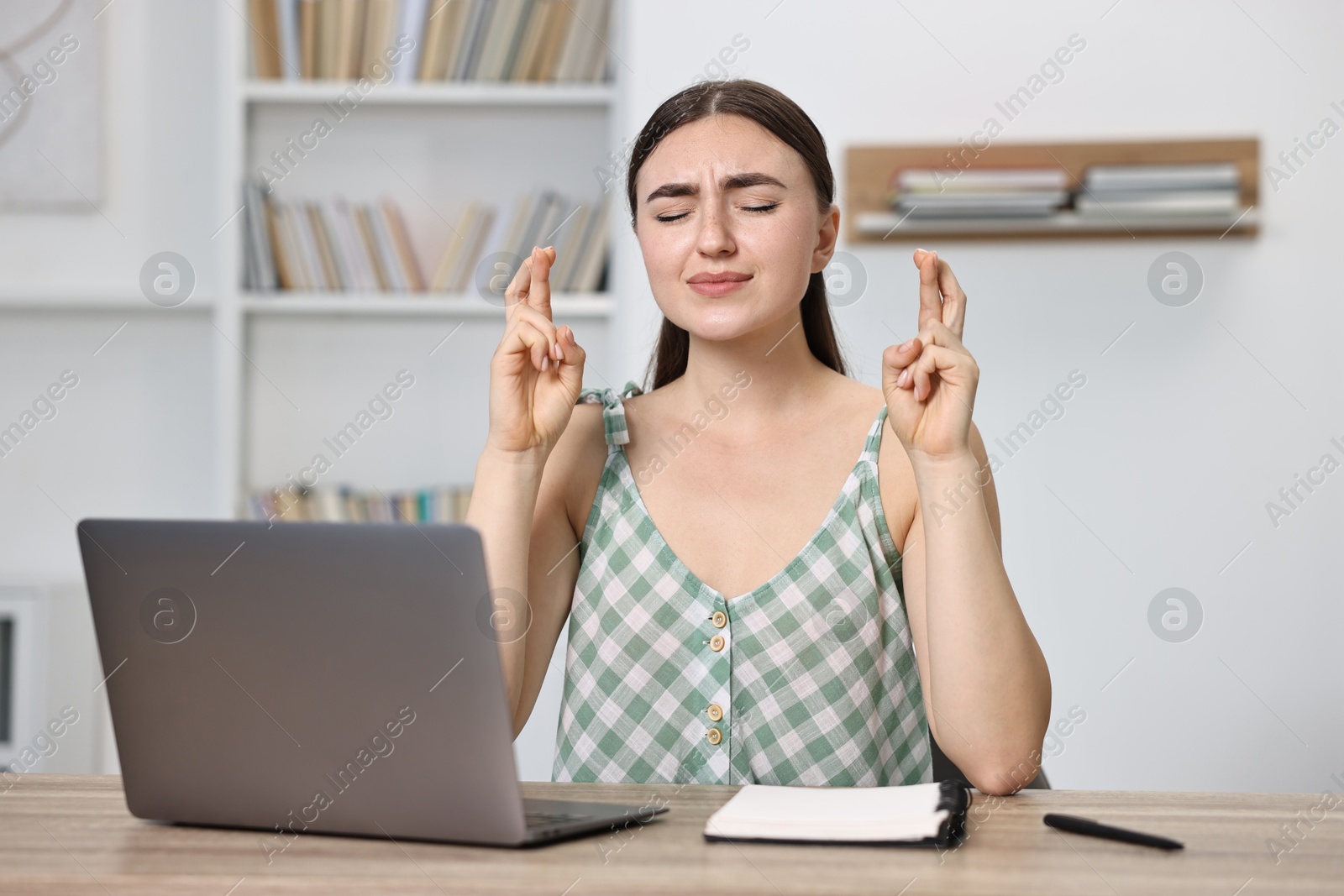 Photo of Worried student with crossed fingers at table with laptop indoors. Hope for good exam result