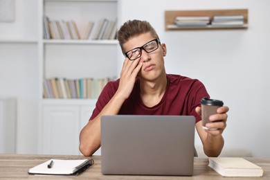 Photo of Tired student preparing for exam with laptop and coffee at table indoors