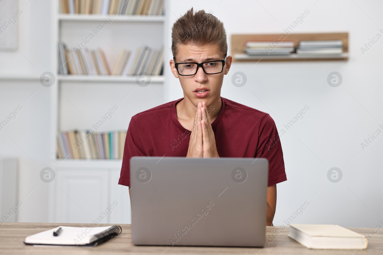 Photo of Worried student praying for good exam result at table with laptop indoors