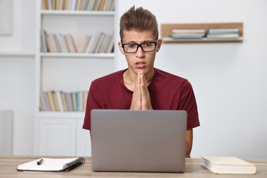 Worried student praying for good exam result at table with laptop indoors