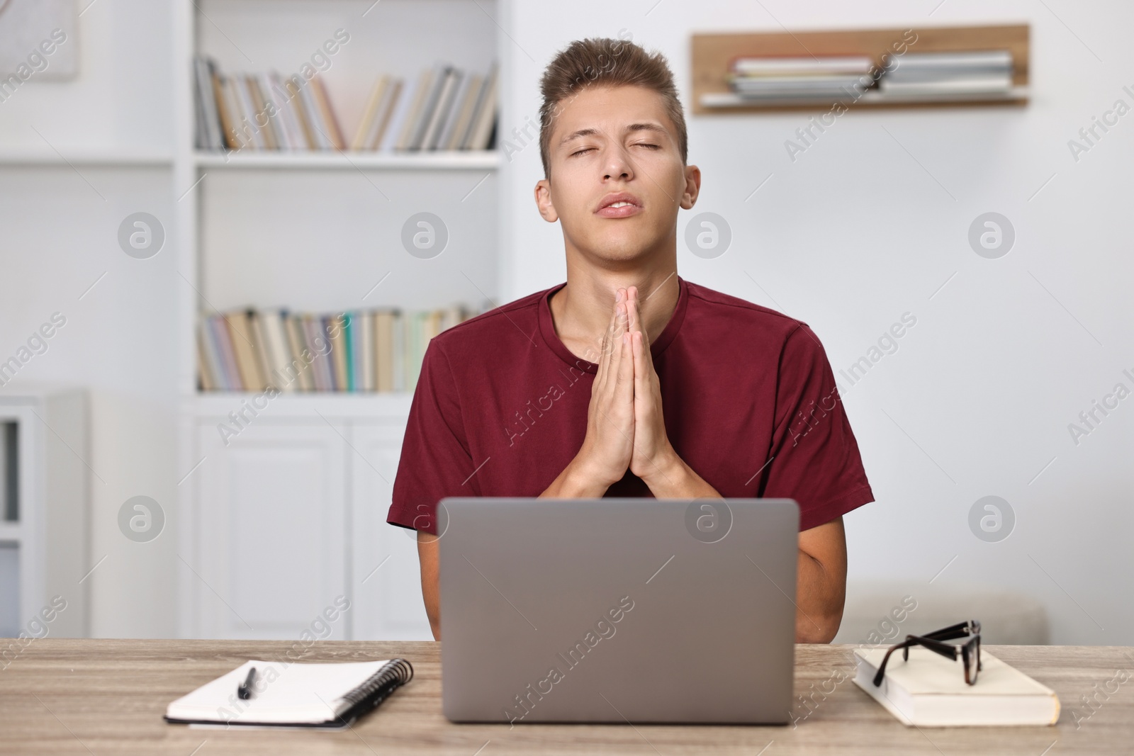 Photo of Worried student praying for good exam result at table with laptop indoors