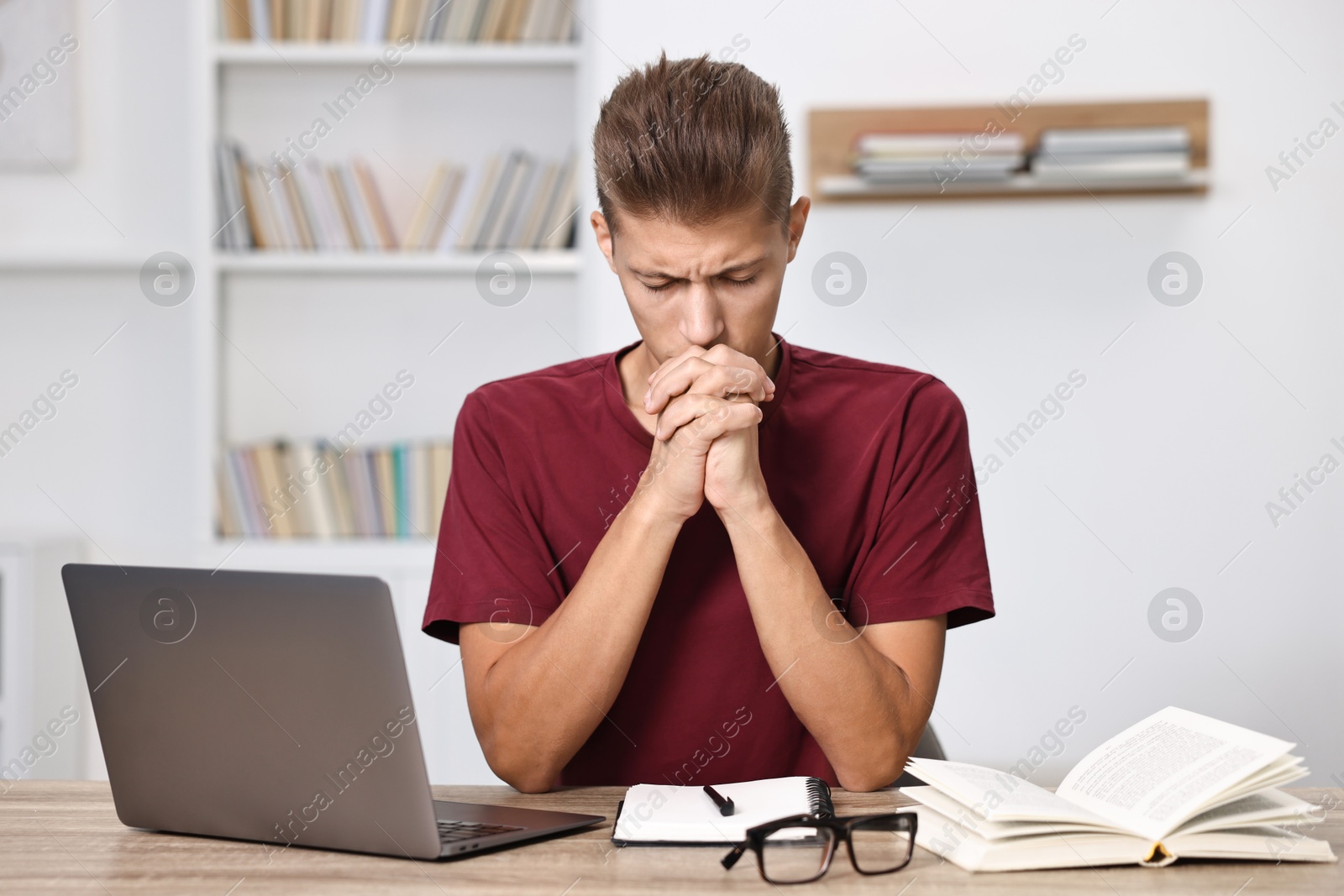 Photo of Worried student praying for good exam result at table with laptop indoors