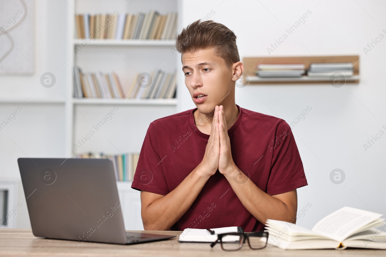 Photo of Worried student praying for good exam result at table with laptop indoors