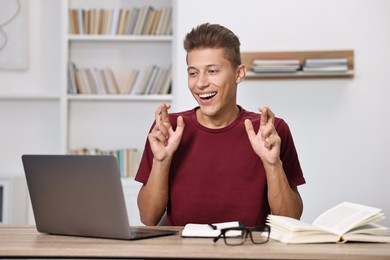 Photo of Student with crossed fingers at table with laptop indoors. Hope for good exam result