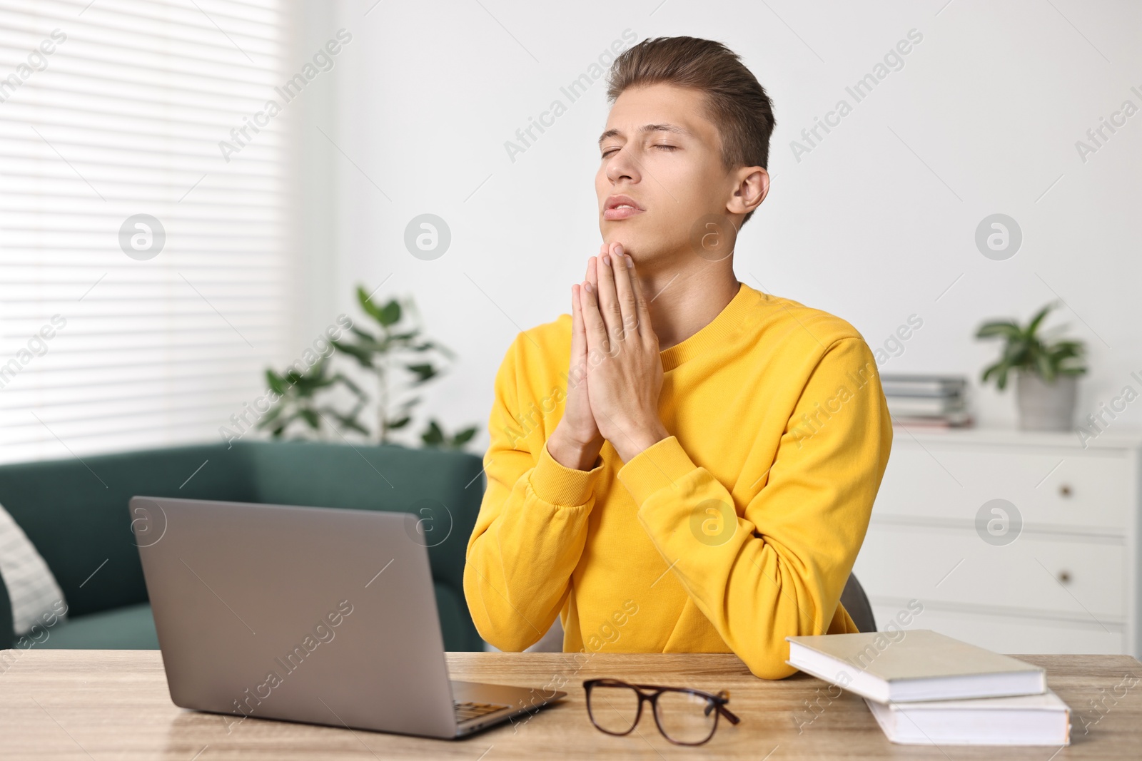 Photo of Student praying for good exam result at table with laptop indoors