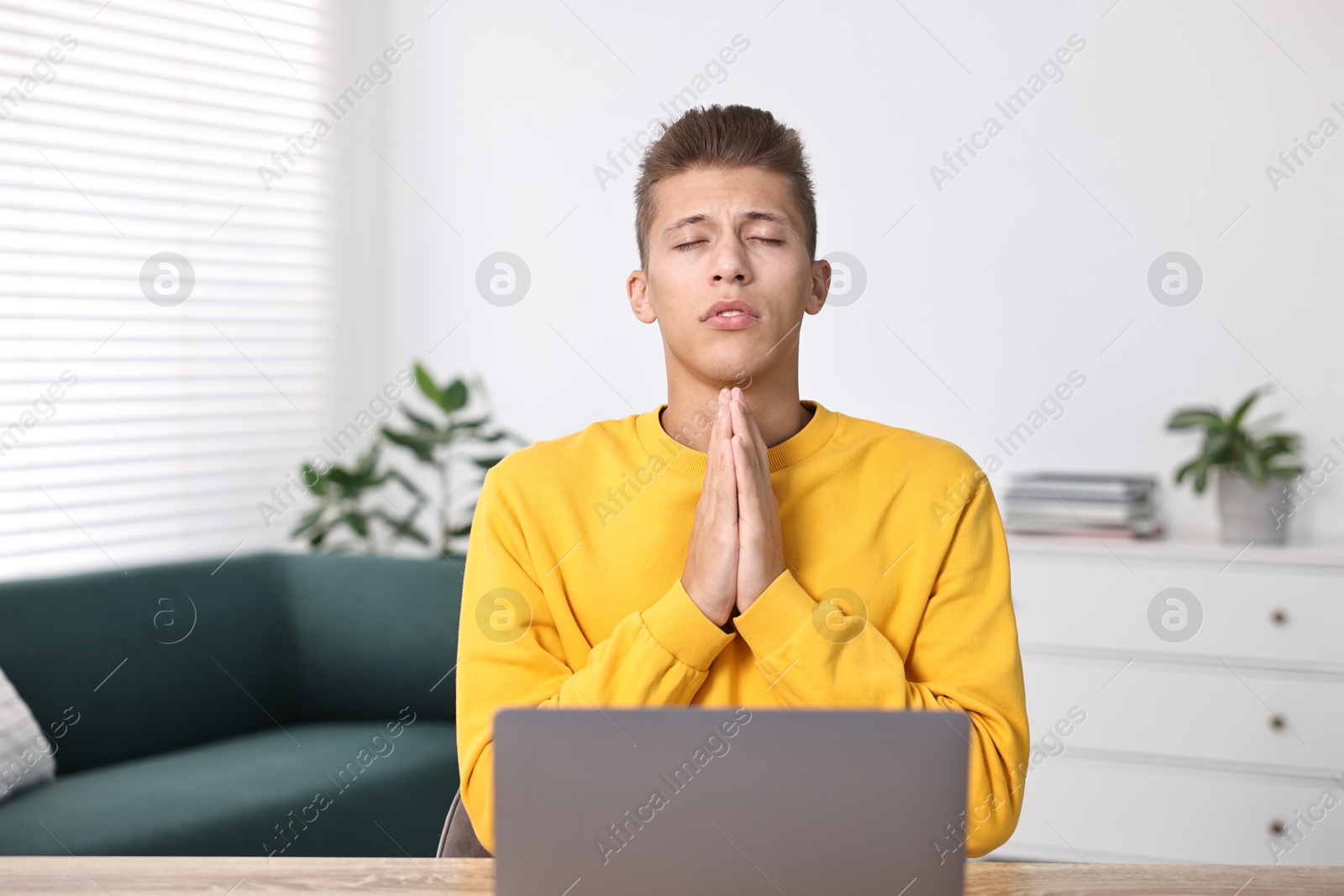 Photo of Student praying for good exam result at table with laptop indoors